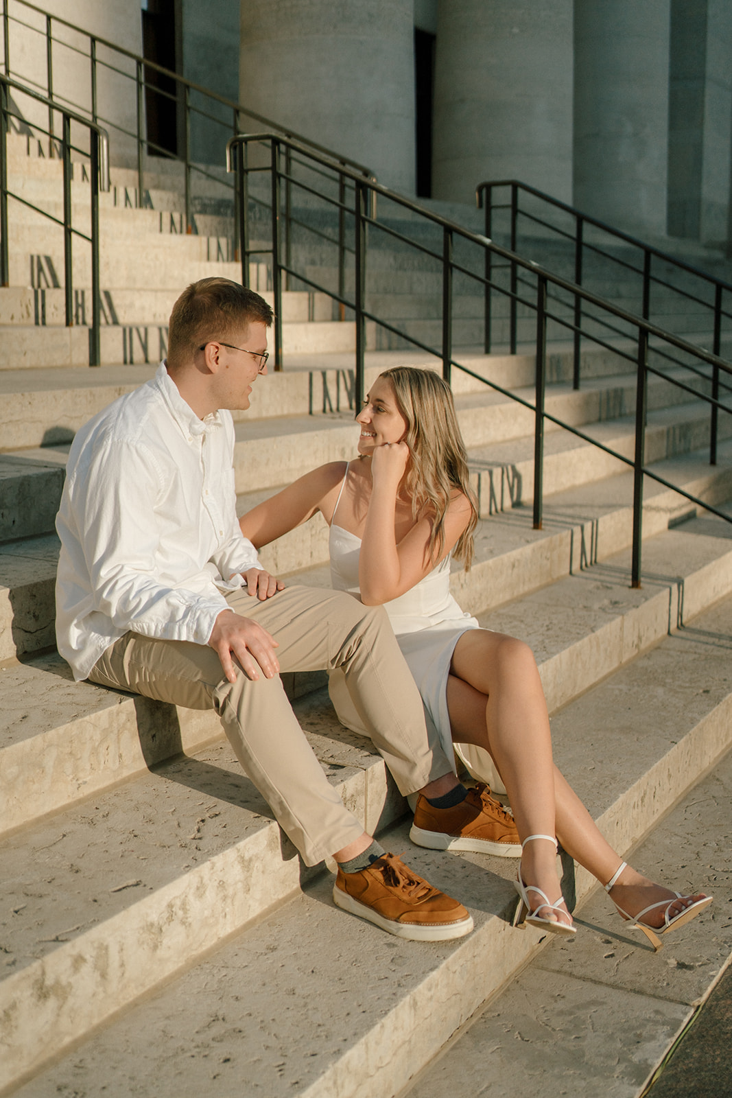 Engaged couple smiles at each other while sitting on the steps of the state house