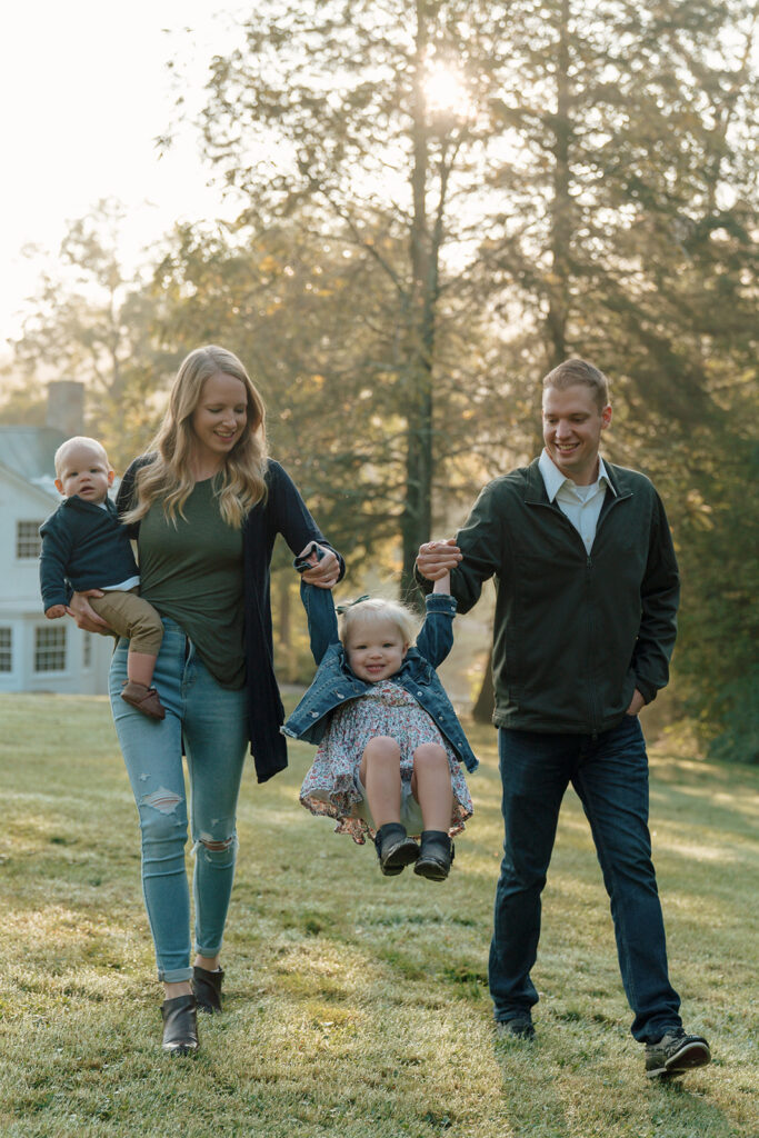 Family of four walk together swinging toddler in between them while mom holds baby on hip