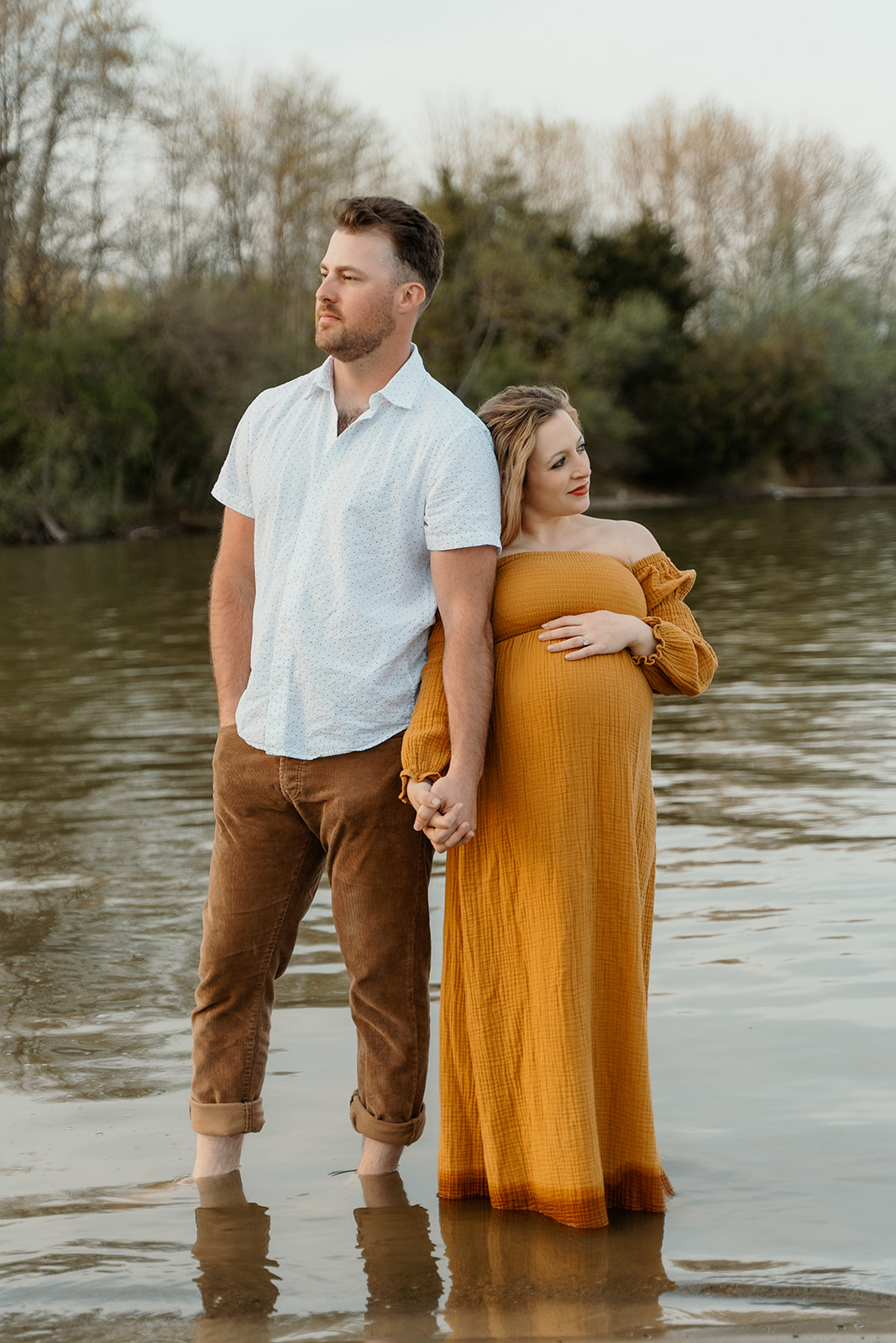 Husband and wife stand ankle deep in the water holding hands during spring maternity session at Alum Creek Beach in Columbus, Ohio