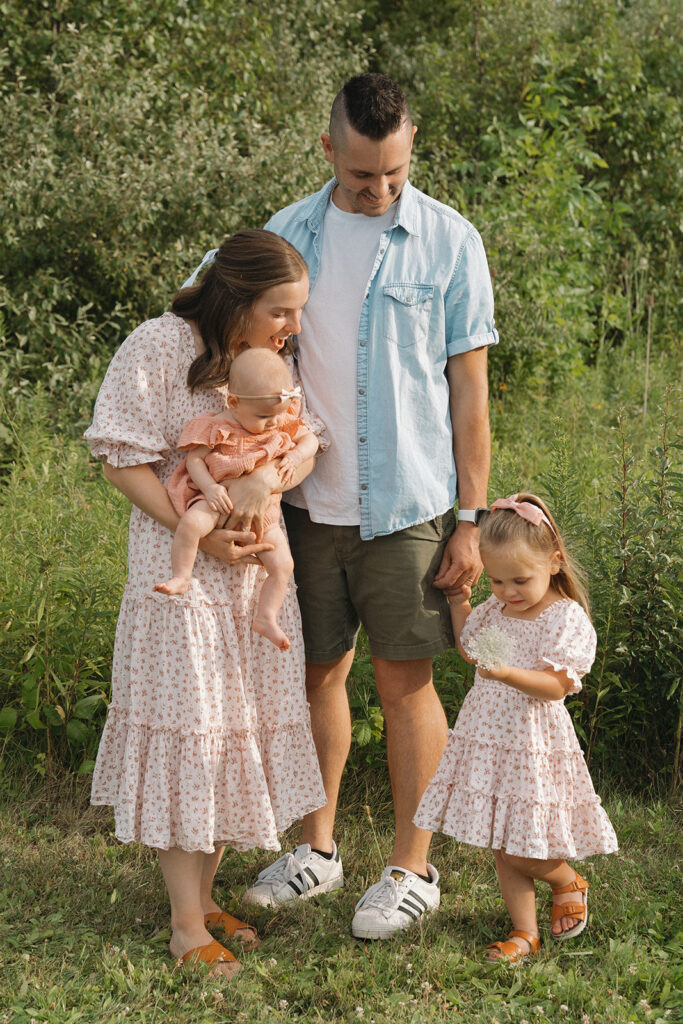 Family of four stands together interacting with toddler who just picked a flower