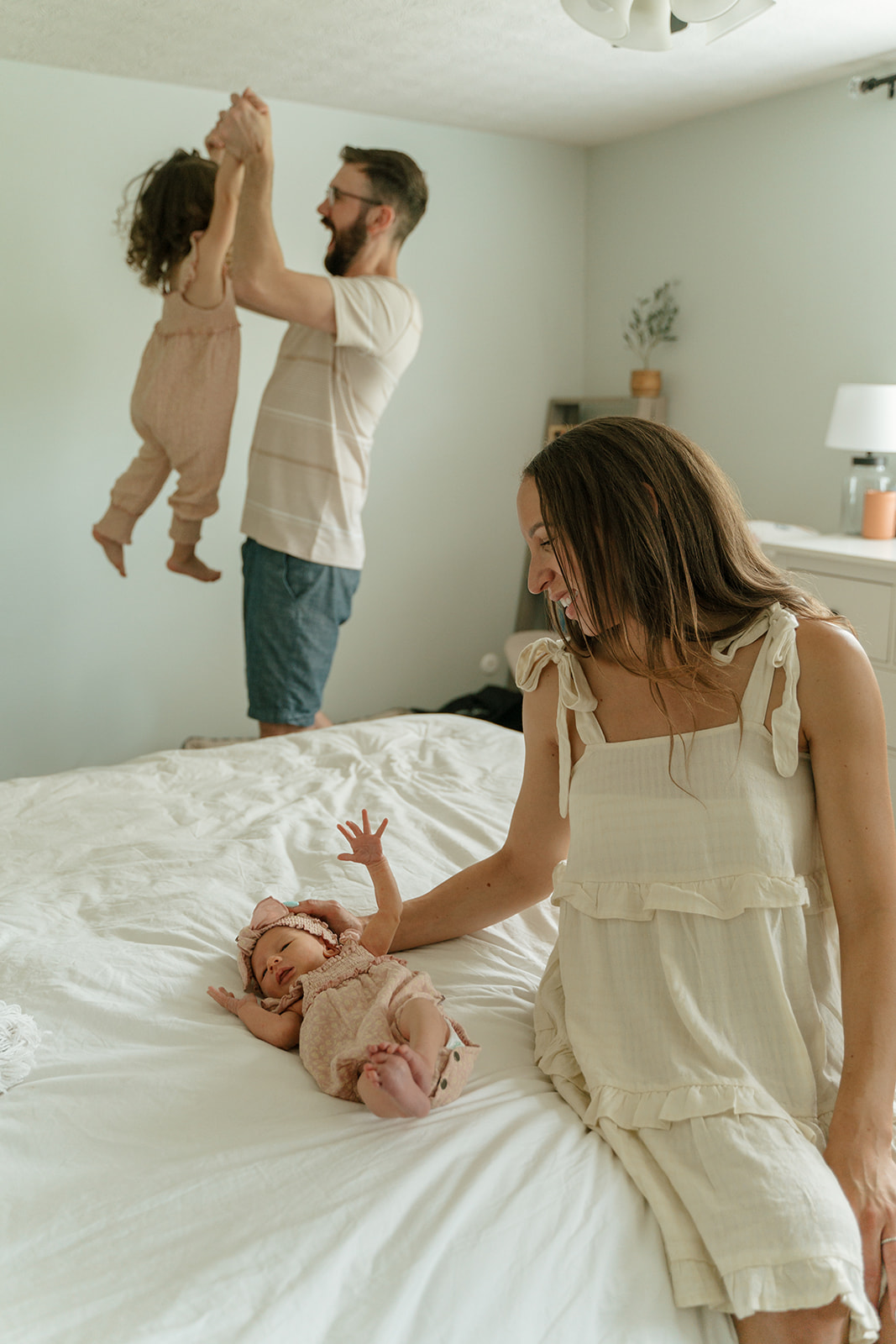 Mother soothes newborn baby while dad plays with toddler in the background during in-home family newborn photoshoot