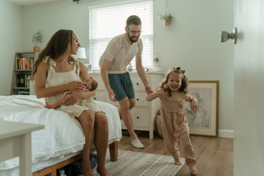 Dad chases toddler around the room while mom watches holding their newborn daughter