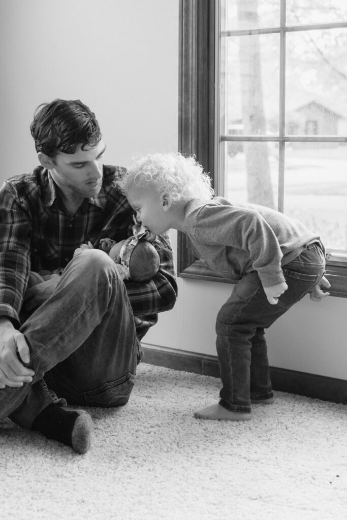 Toddler kisses newborn sister's head while dad holds his new daughter during in home newborn session