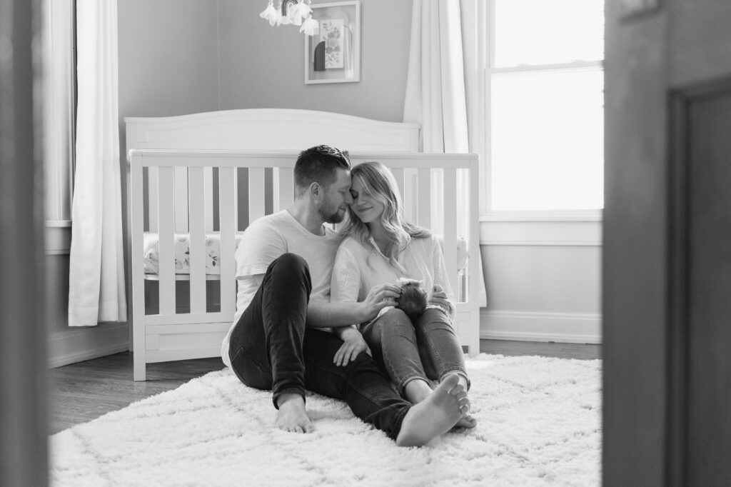 New mom and dad snuggle together in front of crib while holding new baby.